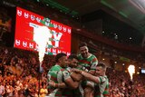 South Sydney players embrace in celebration as a flame shoots up in front of a scoreboard saying 'TRY' in the background.