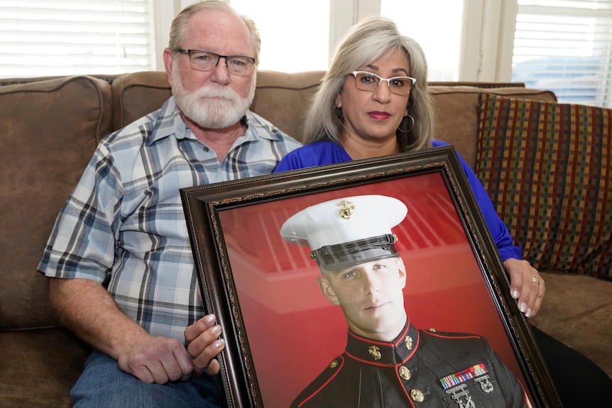 A middle aged man and woman sit on a couch, holding a framed photo of their son, who is in military uniform