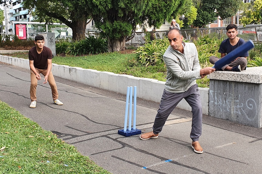 A man swings a cricket bat in the park in fron to of cricket stumps.