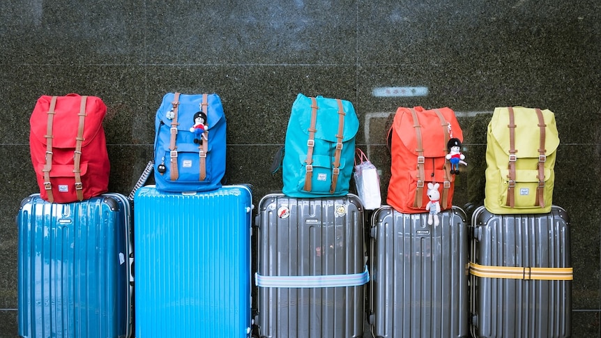 A row of 5 suitcases lined up against a black tiled wall with back packs on top of each.
