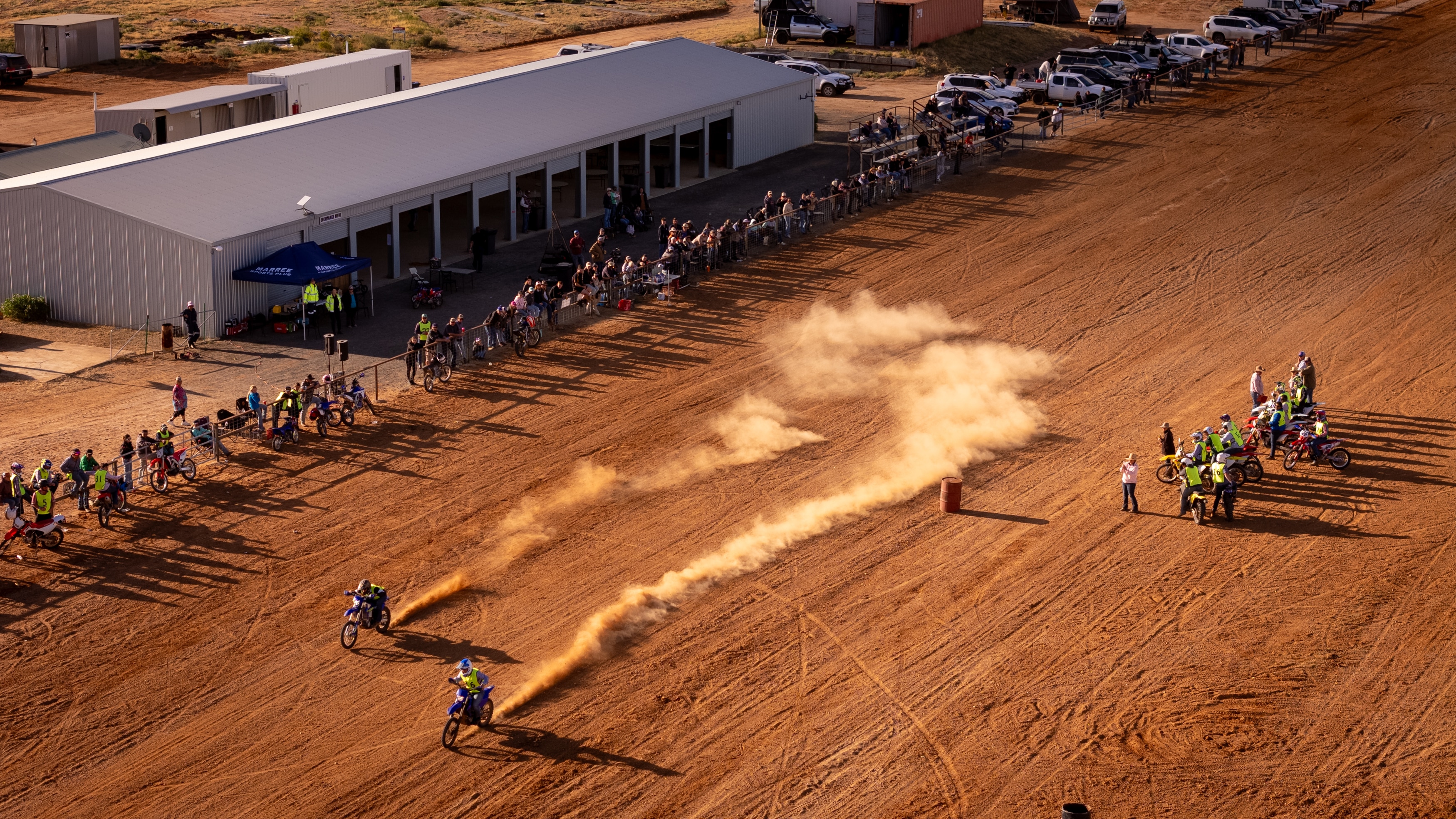 A birds-eye-view of two motorbike on a dirt track, with a crowd of people against a fence watching