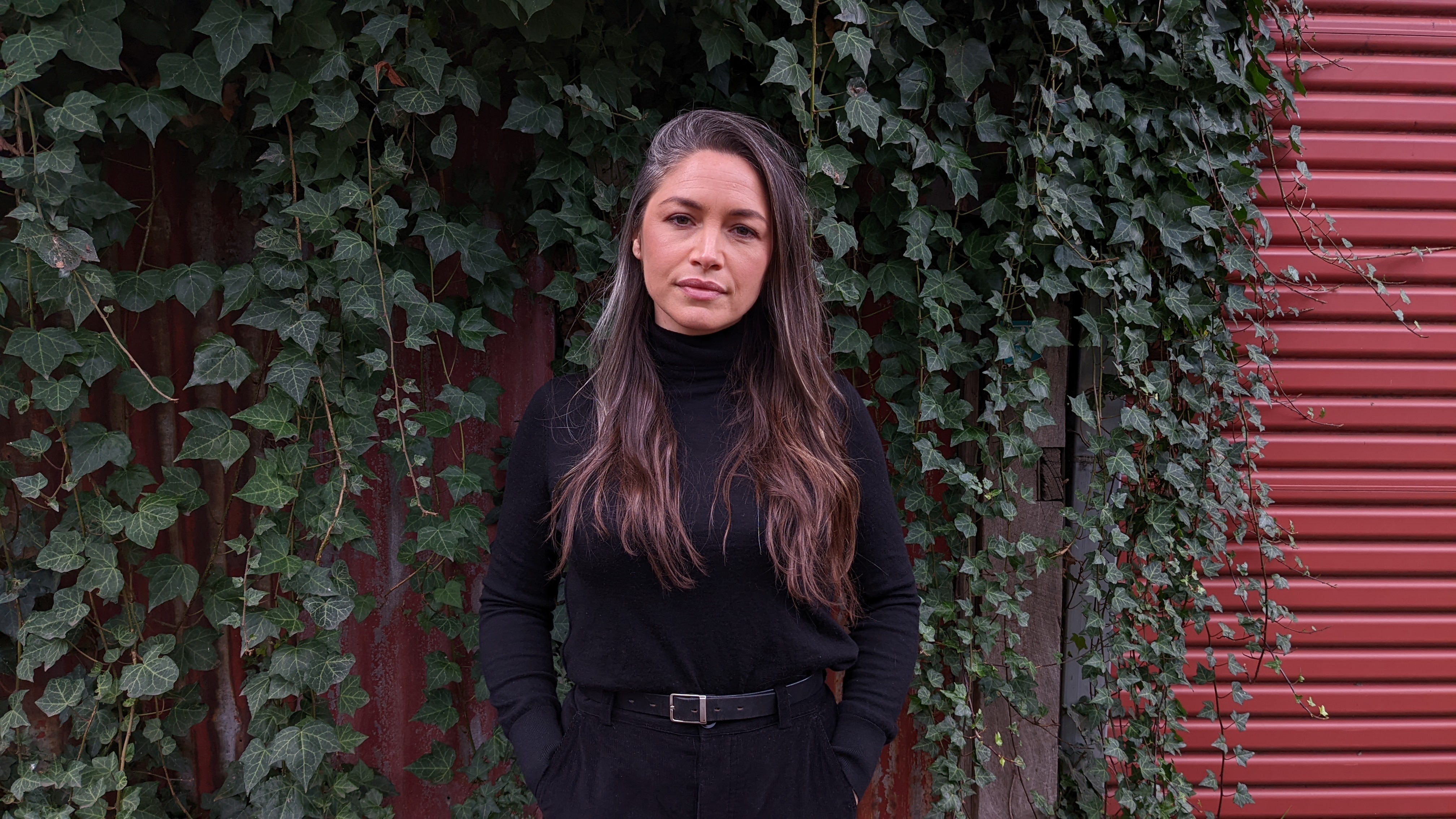 A woman wearing black with long brown hair stands in front of a red garage door covered with hanging green vines. 