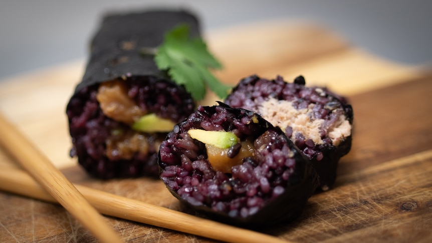Slices of sushi roll filled with dark purple coloured rice, tuna and cucumber on a board with chopsticks and garnish.