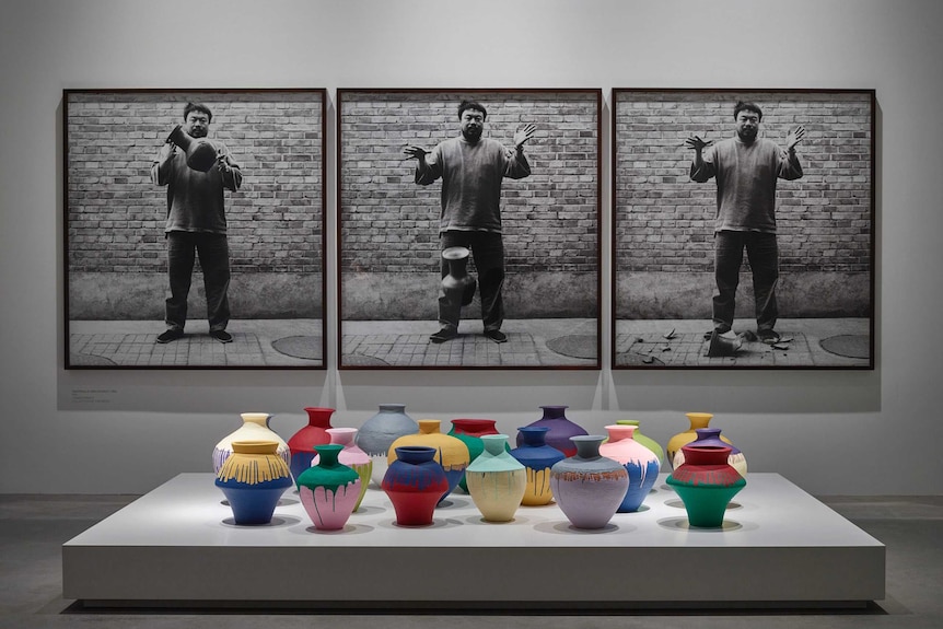 An art installation of a a collection of ceramic pots in front of a series of three photographs showing a man smashing a pot.