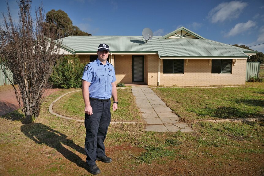 A policeman stands in front of a house with light-coloured bricks, steel roof, green trimmings, a bush behind him.