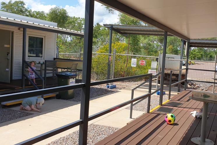 Two kids play in front of a low-set worker's cabin with a wooden balcony, wire fencing is seen surrounding the area.