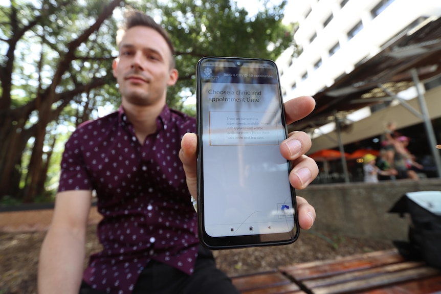 A young man holds his phone close to the camera. A government message can be seen saying 'no bookings available'.