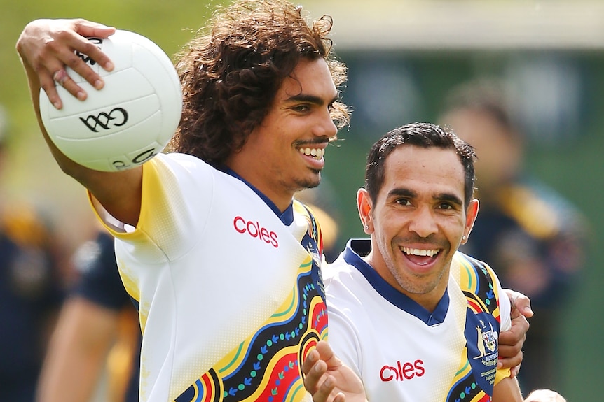 Tony Armstrong tiene su brazo alrededor de Eddie Betts mientras sostiene una pelota redonda.  Ambos están sonriendo