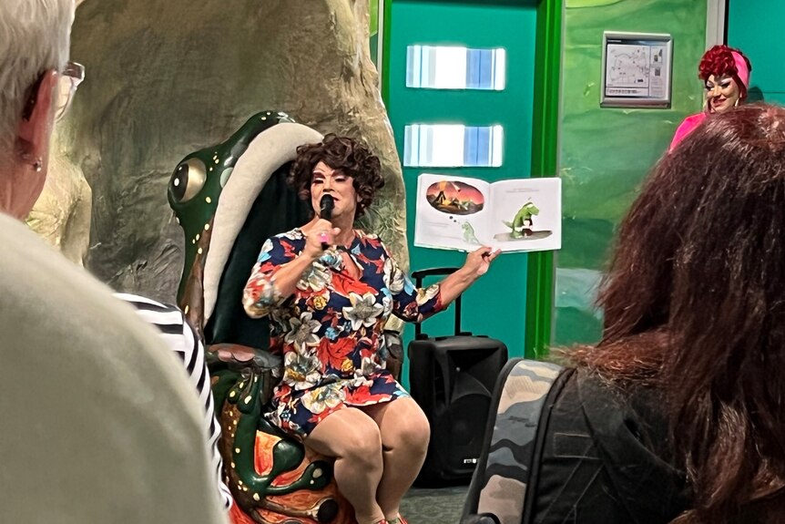 A person wearing a dress holding a microphone and sitting in a frog-shaped chair showing a book