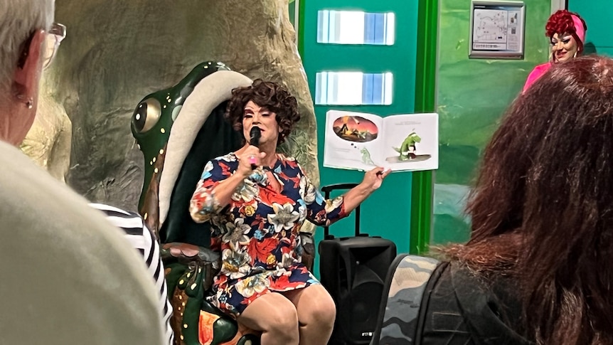 A person wearing a dress holding a microphone and sitting in a frog-shaped chair showing a book.