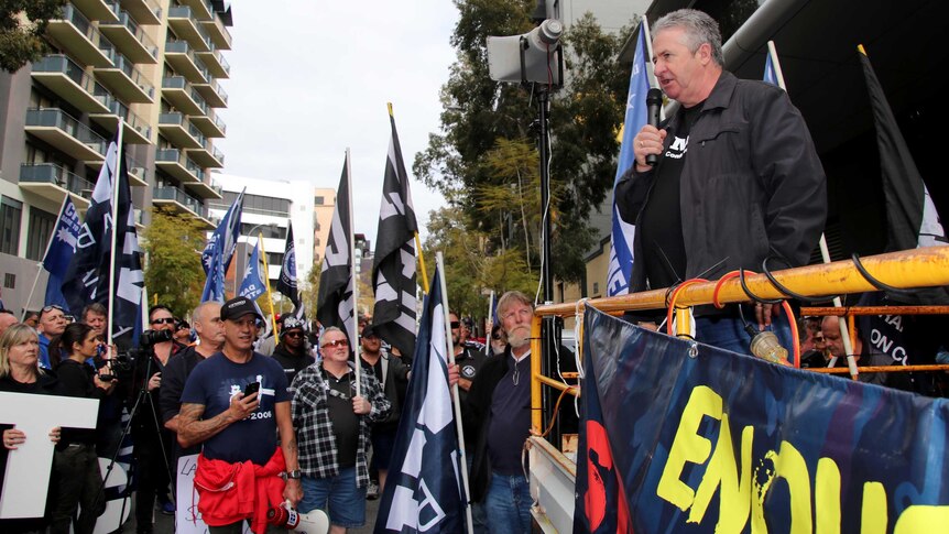 Man stands on back of a ute speaking into a microphone to a crowd of flag holding supporters.