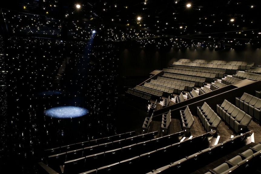 Rows of empty seats as thousands of spotlights glimmer overhead. A spotlight shines a circle on the stage below.