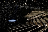 Rows of empty seats as thousands of spotlights glimmer overhead. A spotlight shines a circle on the stage below.
