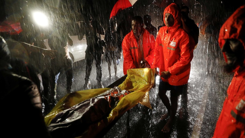Men in bright orange jackets carry a body in heavy rain.