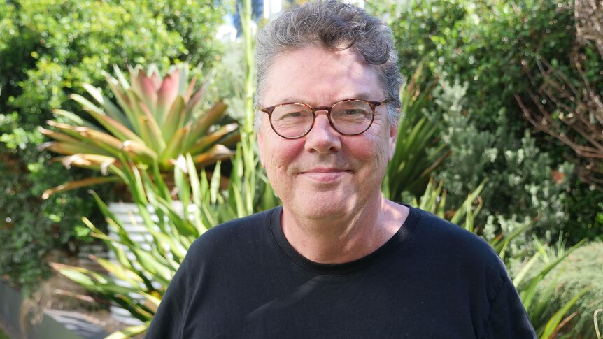 A man in a black t-shirt with green plants behind him. 