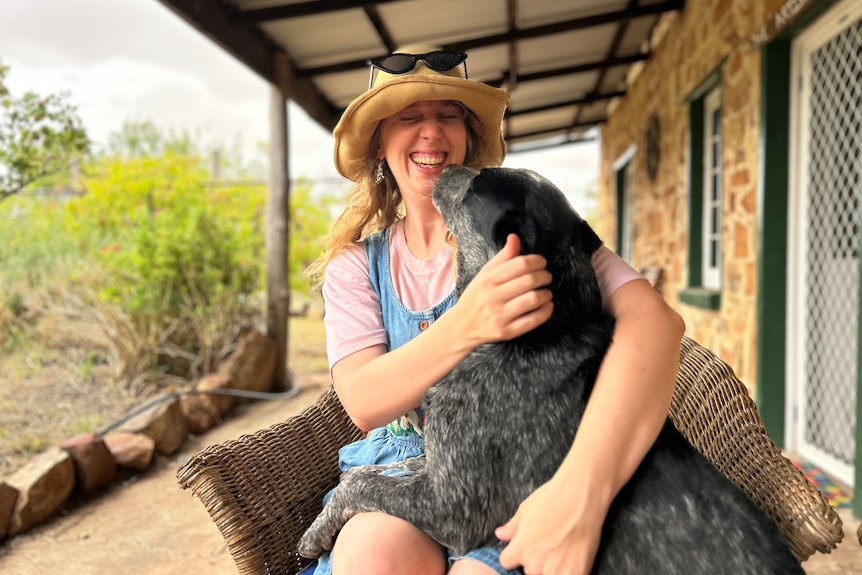 A woman sitting on a wicker chair on a veranda, laughing as a dog licks her face.