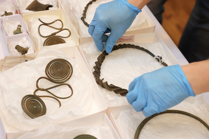 A pair of gloved hands handle an ancient bronze necklace sitting in a white paper box. 