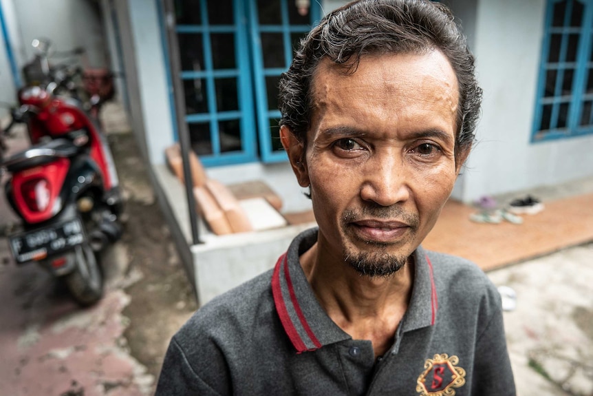 An Indonesian man standing outside his small house with blue shutters