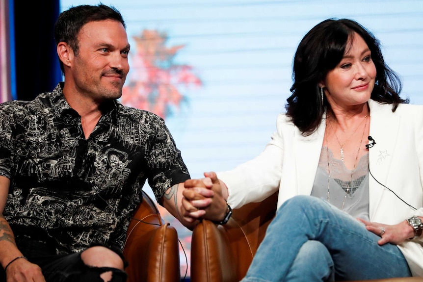 A man in a black patterned shirt and a woman in a white jacket sit in chairs holding hands.