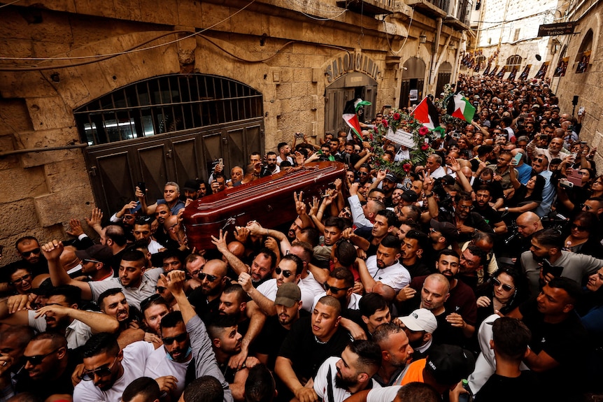 Thousands attend funeral procession along street carrying a coffin.