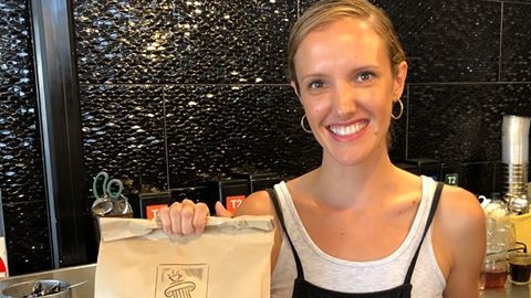A smiling blonde woman holds a brown paper food bag stamped Roma Bar and a lidded takeaway coffee cup.
