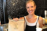A smiling blonde woman holds a brown paper food bag stamped Roma Bar and a lidded takeaway coffee cup.