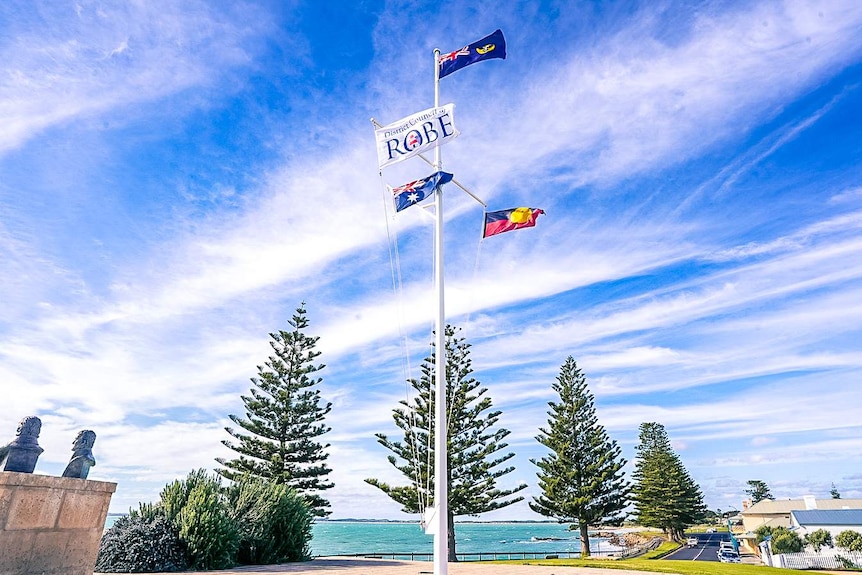 A large flag pole with the Australian, Aboriginal and Robe District Council flags. Large pine trees and an aqua coastline behind