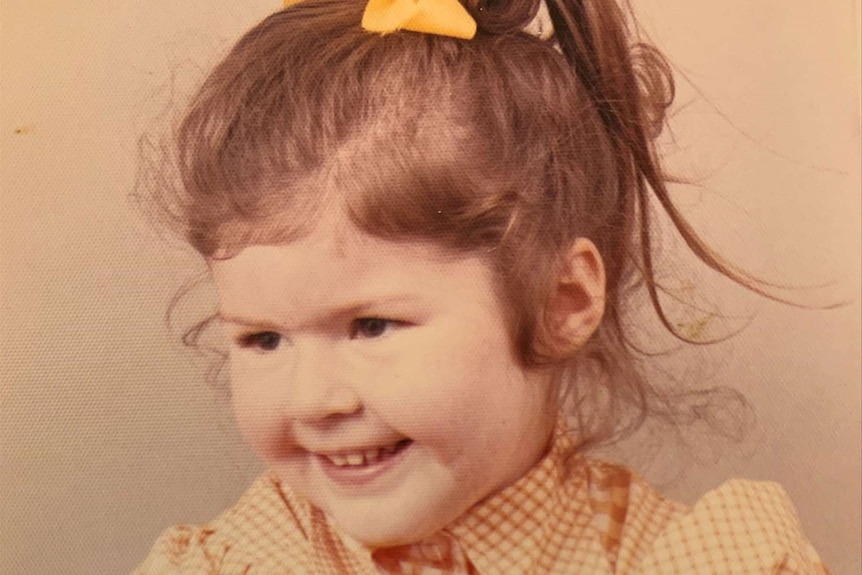 A young girl poses for a photo wearing a yellow and white gingham dress with her hair in a ponytail.