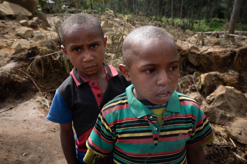 Two young PNG boys stand in front of a pile of rocks, one looking directly at the camera.