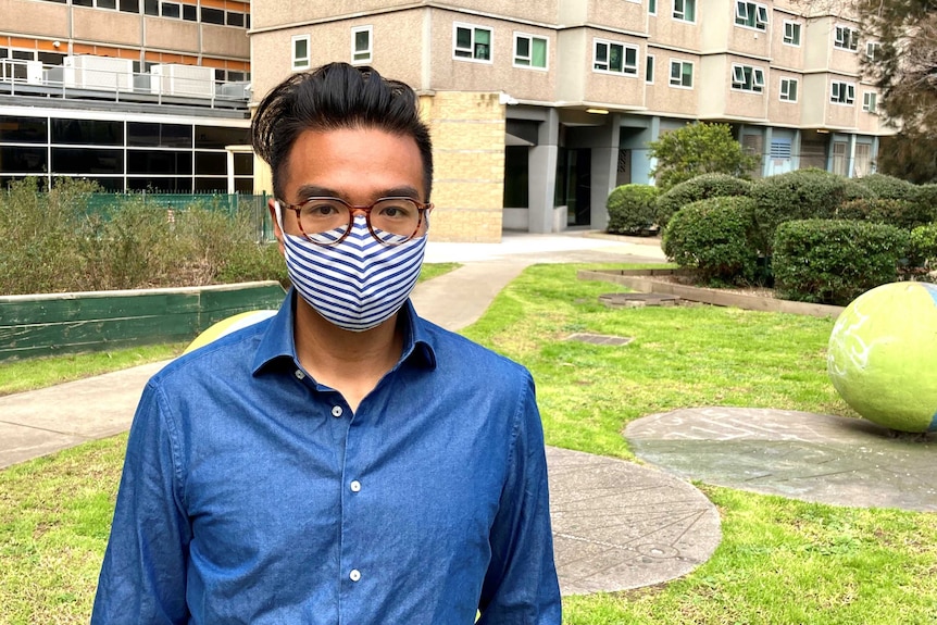 Lawyer Daniel Nguyen standing in front of a residential tower block on Melbourne, wearing glasses, a blue and white striped mask