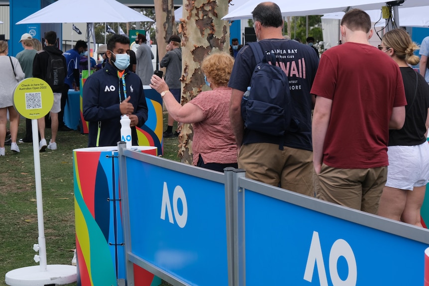 A masked Australian Open staff looks at a phone being held up by a woman at the front of the queue, checking vaccination status.
