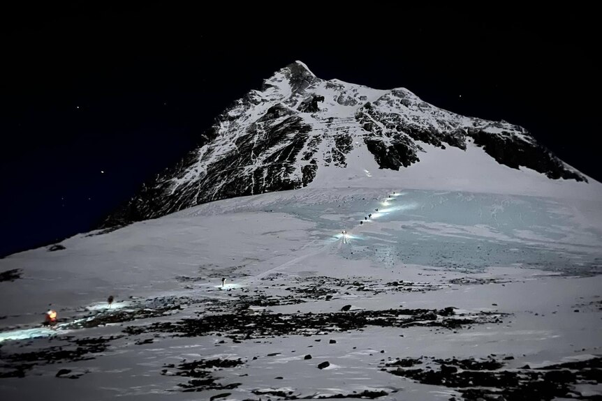 A line of climbers make their way up to the peak of Mount Everest in the dark, with a row of lights marking the way.
