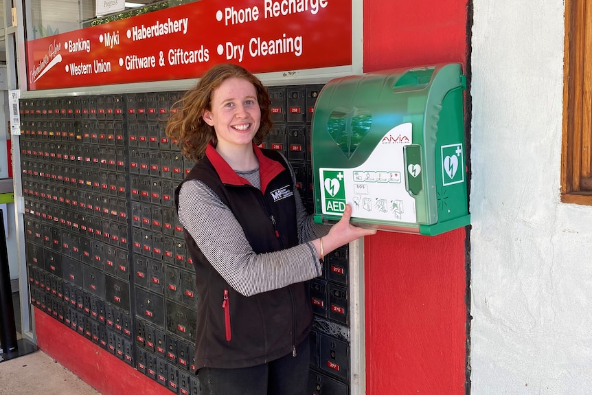 Girl stands next to green defibrillator mounted on a red wall