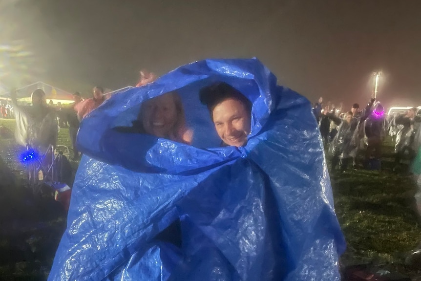 Two people with a tarp wrapped around them at a concert outside.