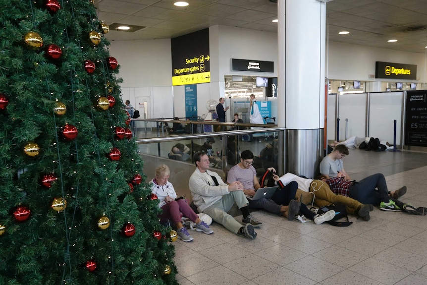 A line of people sitting on the ground of an airport. Some are charging electronic devices, others appear to be sleeping.
