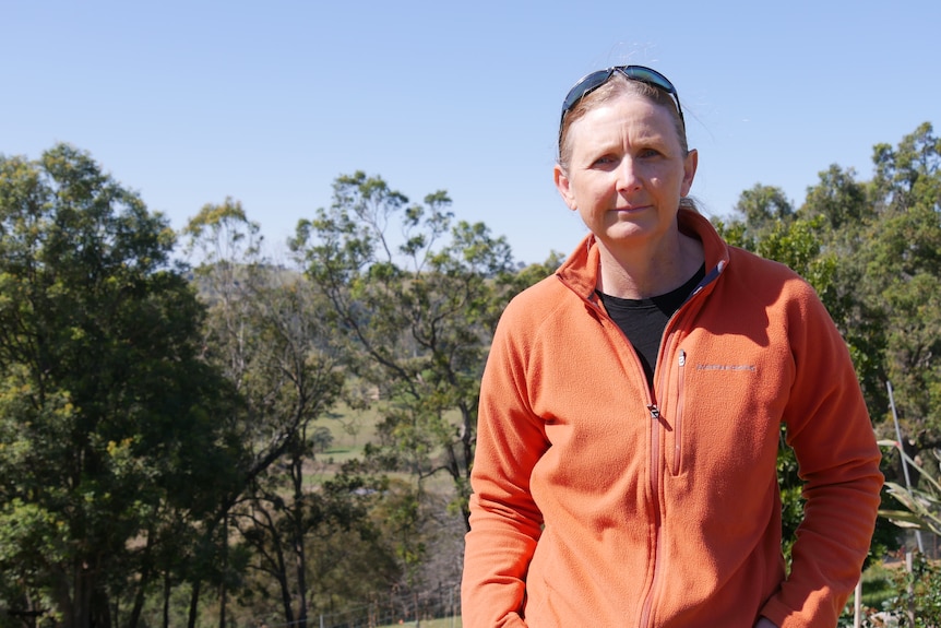 a woman stands with an orange jumper with a view of trees in the background 