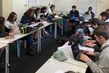 Students sitting at desks in a narrow classroom, laptops and bags cover the table surface