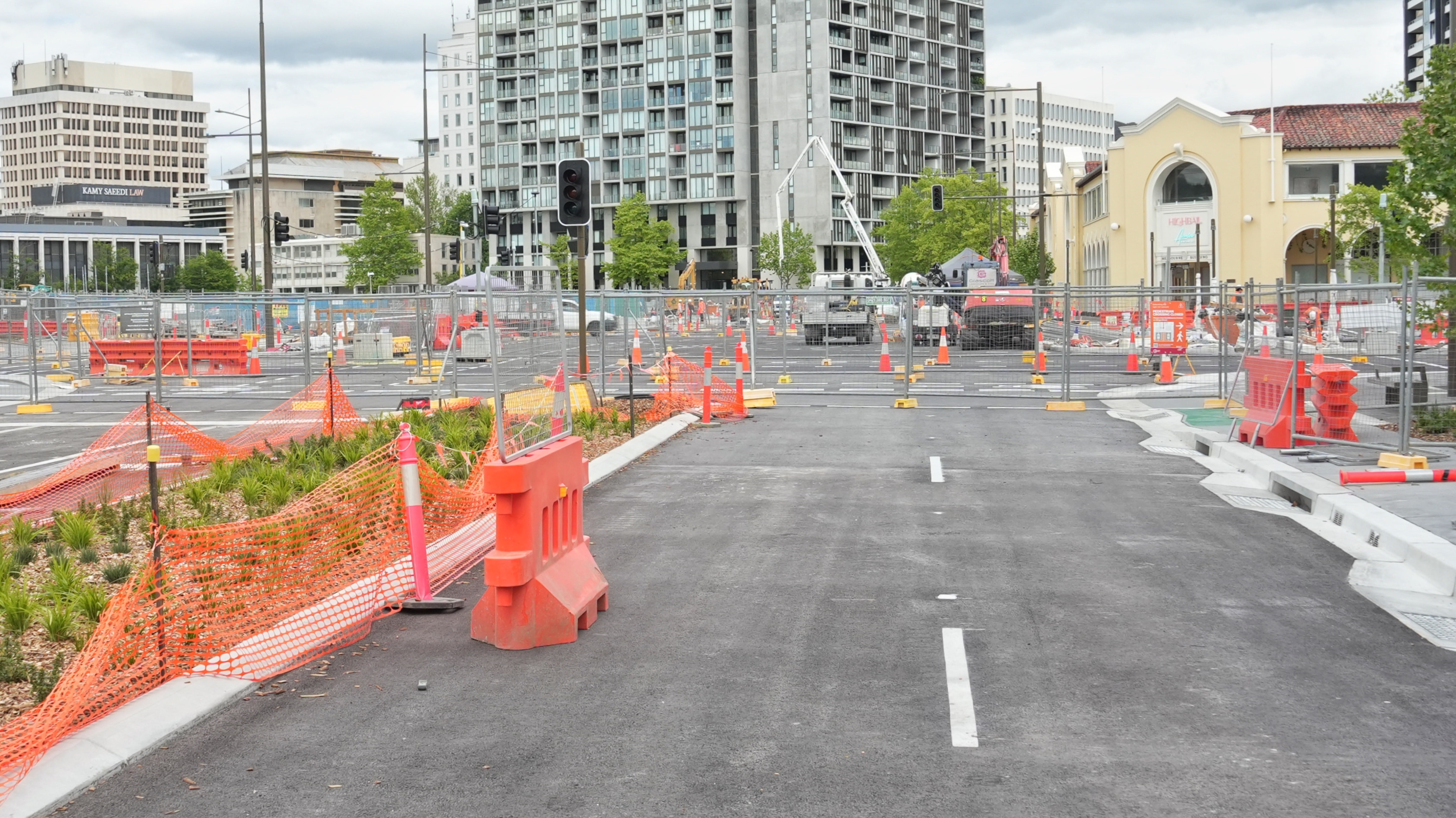 Road blocked off by fences and orange road blocks. A crane in the distance and buildings in the distance.