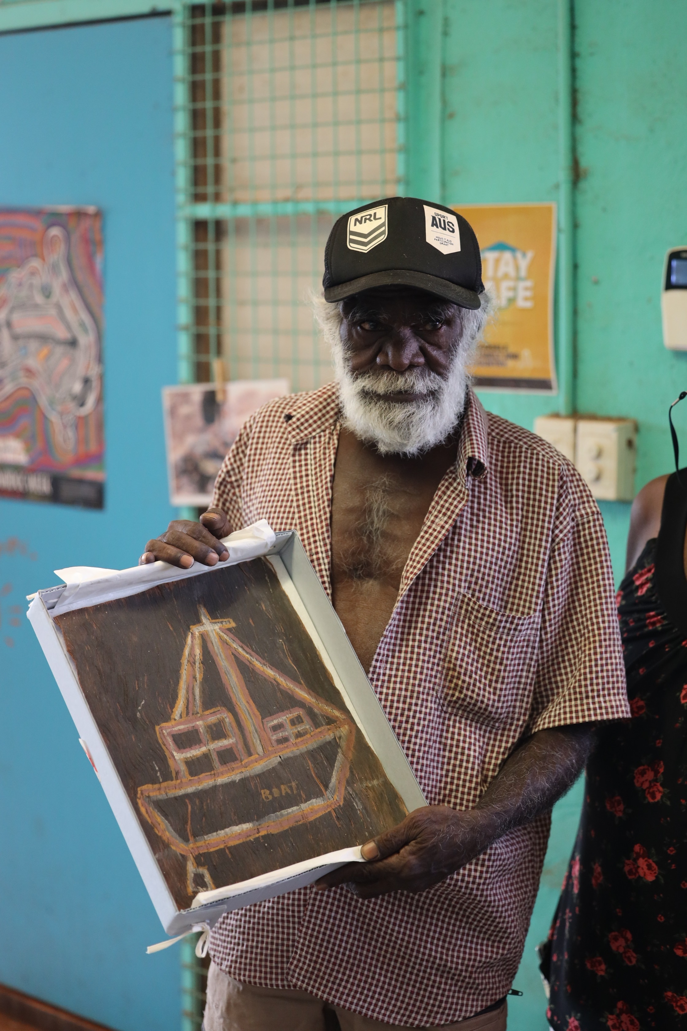 An Aboriginal man wearing a cap holds a small painting