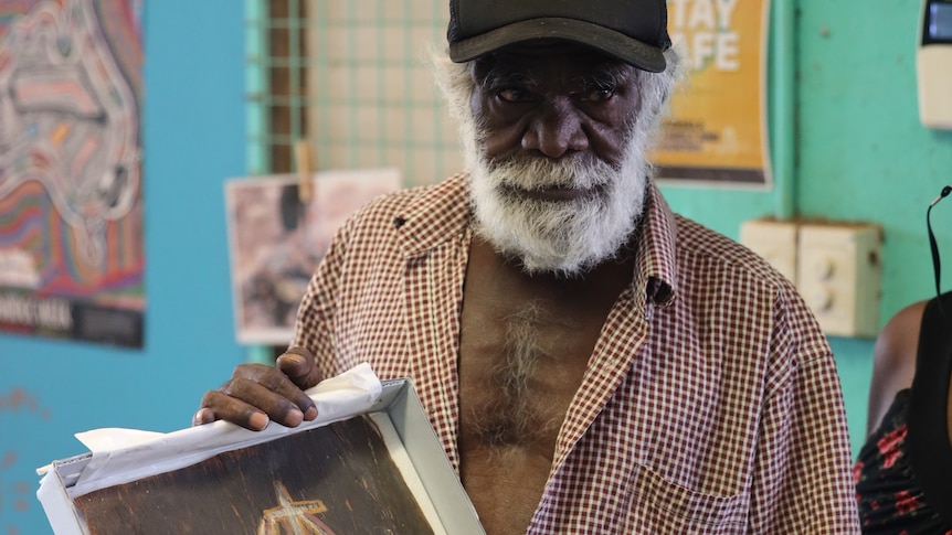 An Aboriginal man wearing a cap holds a small painting