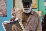 An Aboriginal man wearing a cap holds a small painting