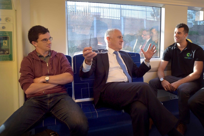 Front on view of Malcolm Turnbull sitting between two passengers on a train and gesturing as he talks to one of them.