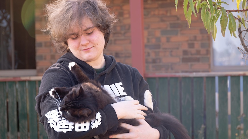A white man with light brown hair stands outside and holds his cat and looks lovingly at it.