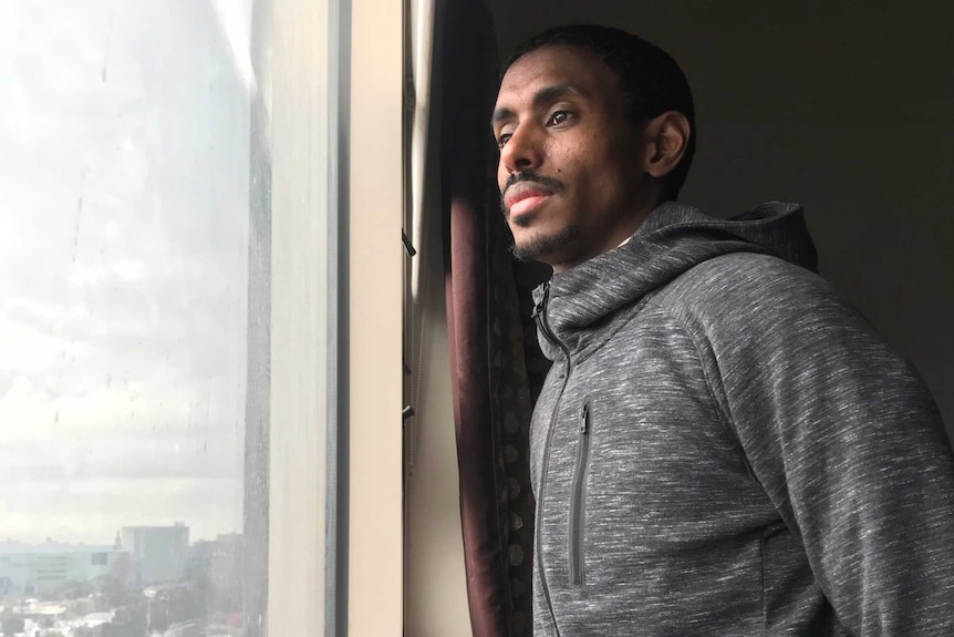 Ahmed Dini, wearing a grey hoodie speckled with white, looking out of the window of the tower block he lives in.