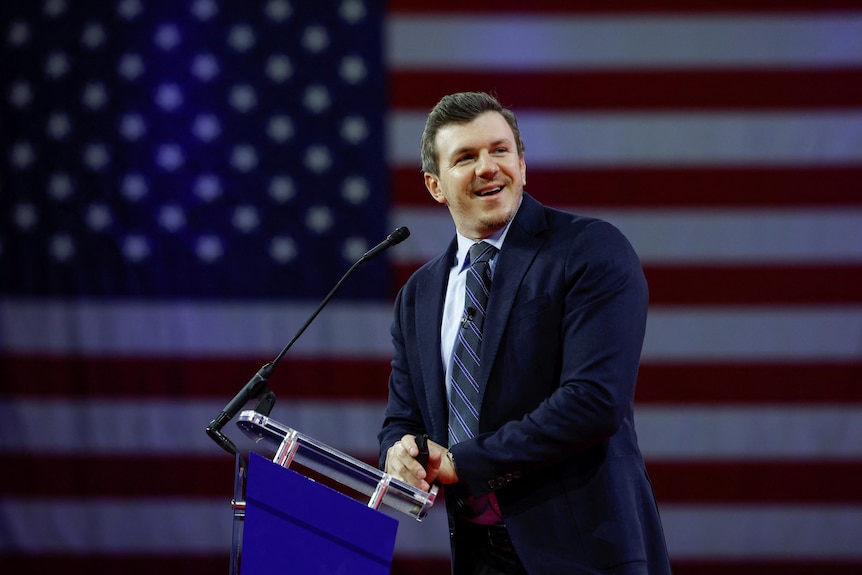 A man stands at a podium speaking against a US flag backdrop 