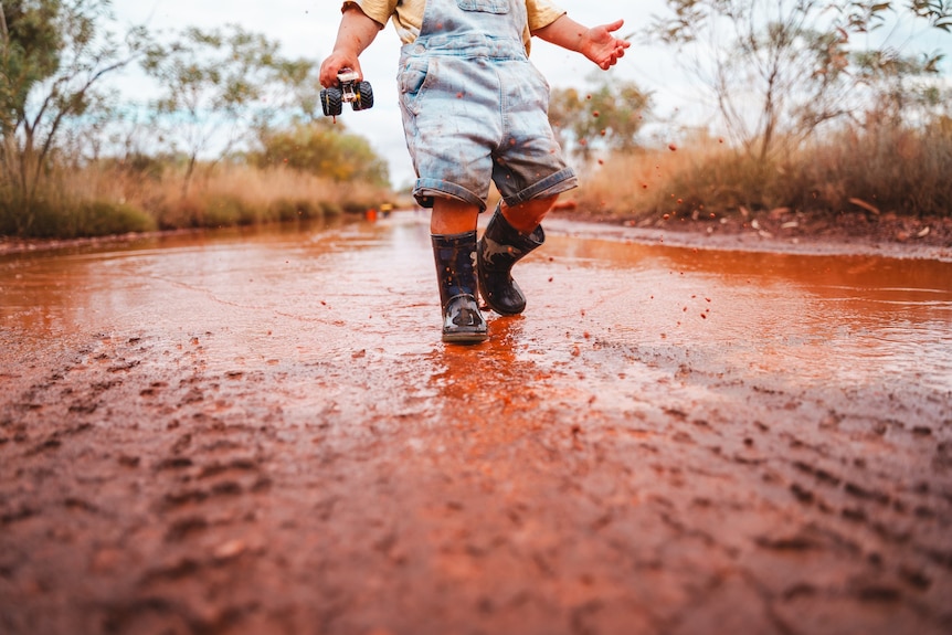 A child in gumboots walks a muddy road holding a toy truck