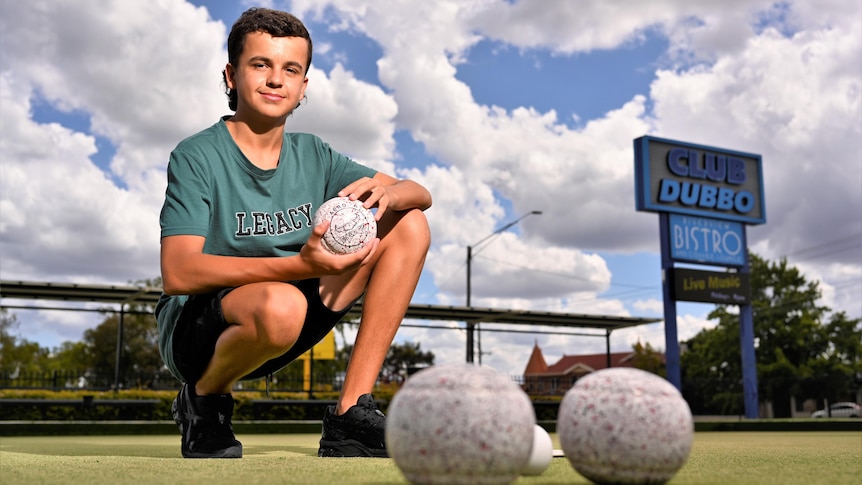A smiling teenager with dark hair kneels down on a bowling green, holding a ball.