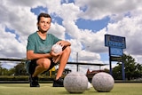 A smiling teenager with dark hair kneels down on a bowling green, holding a ball.