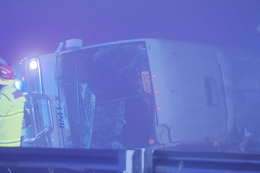 A dark, blue-lit photo shows a bus lying on its side and an emergency services worker.