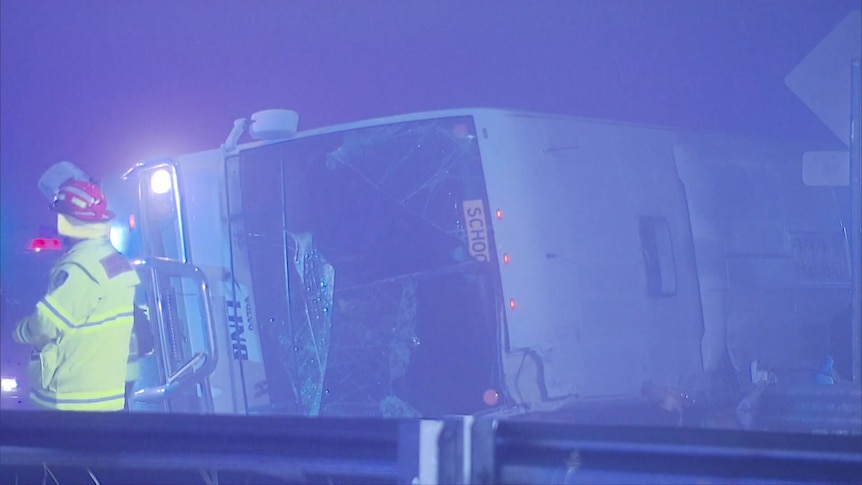 A dark, blue-lit photo shows a bus lying on its side and an emergency services worker.